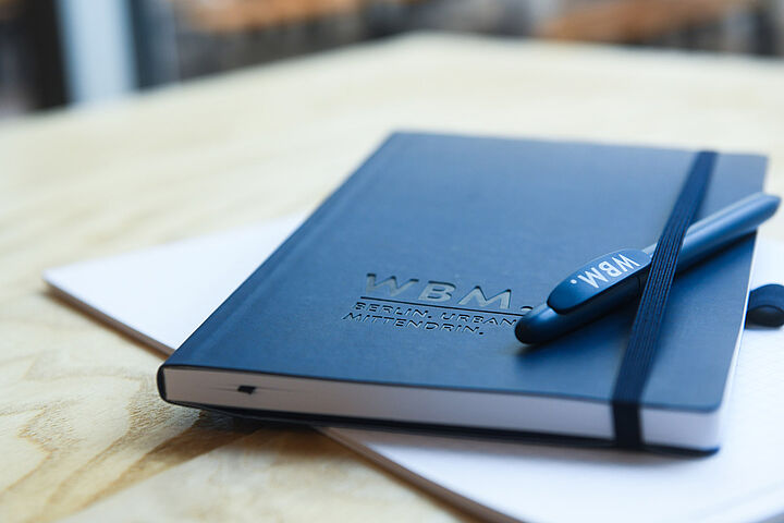 Eine dunkelblaue WBM-Kladde mit einem Gummizug, in den ein Stift geklemmt ist, liegt auf einem Holztisch.